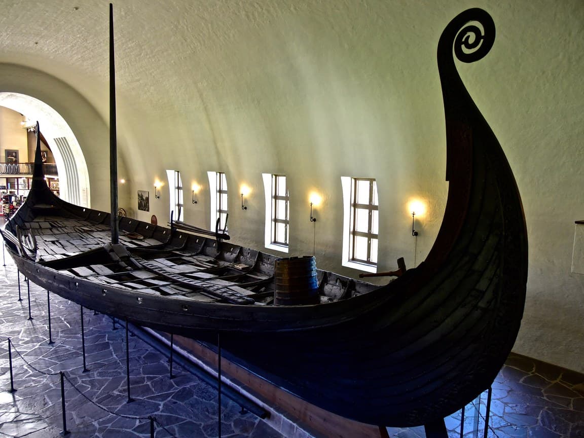 The Oseberg ship in the Viking Ship Museum in Oslo, a dark, clinker-built wooden ship with a tall prow carved in a spiral, seen from the bow.