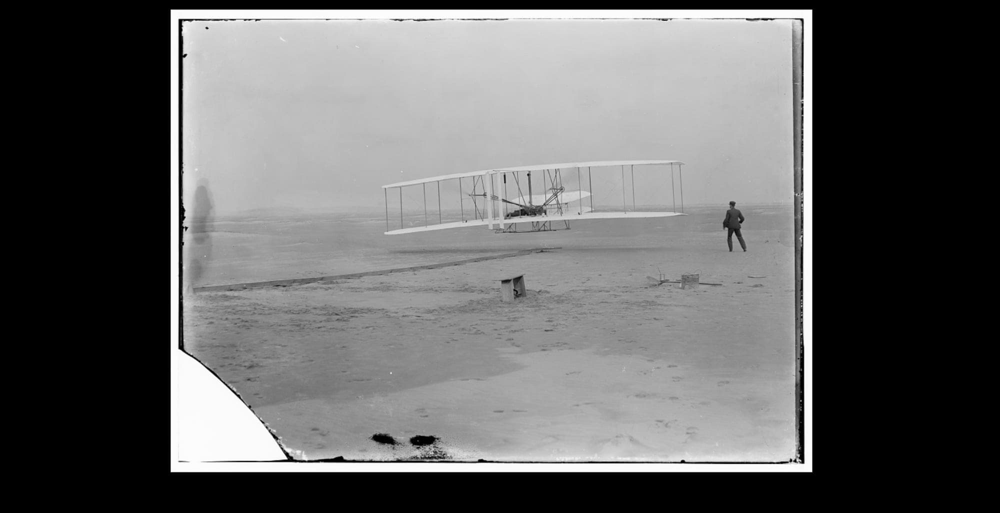The Wright Flyer just after leaving the rail at Kill Devil Hills, Wilbur Wright running alongside.