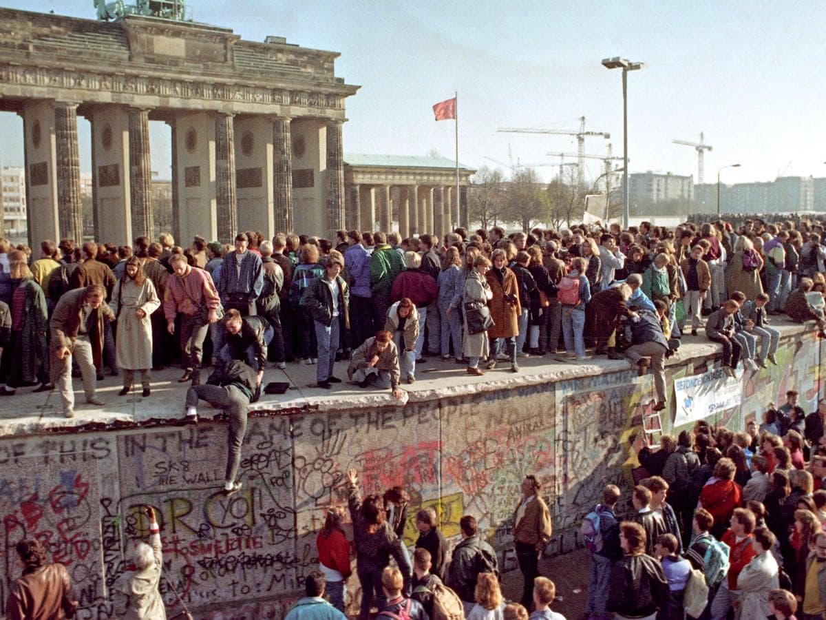 Crowds gathered on top of the Berlin Wall and in the street below, with the Brandenburg Gate behind them.