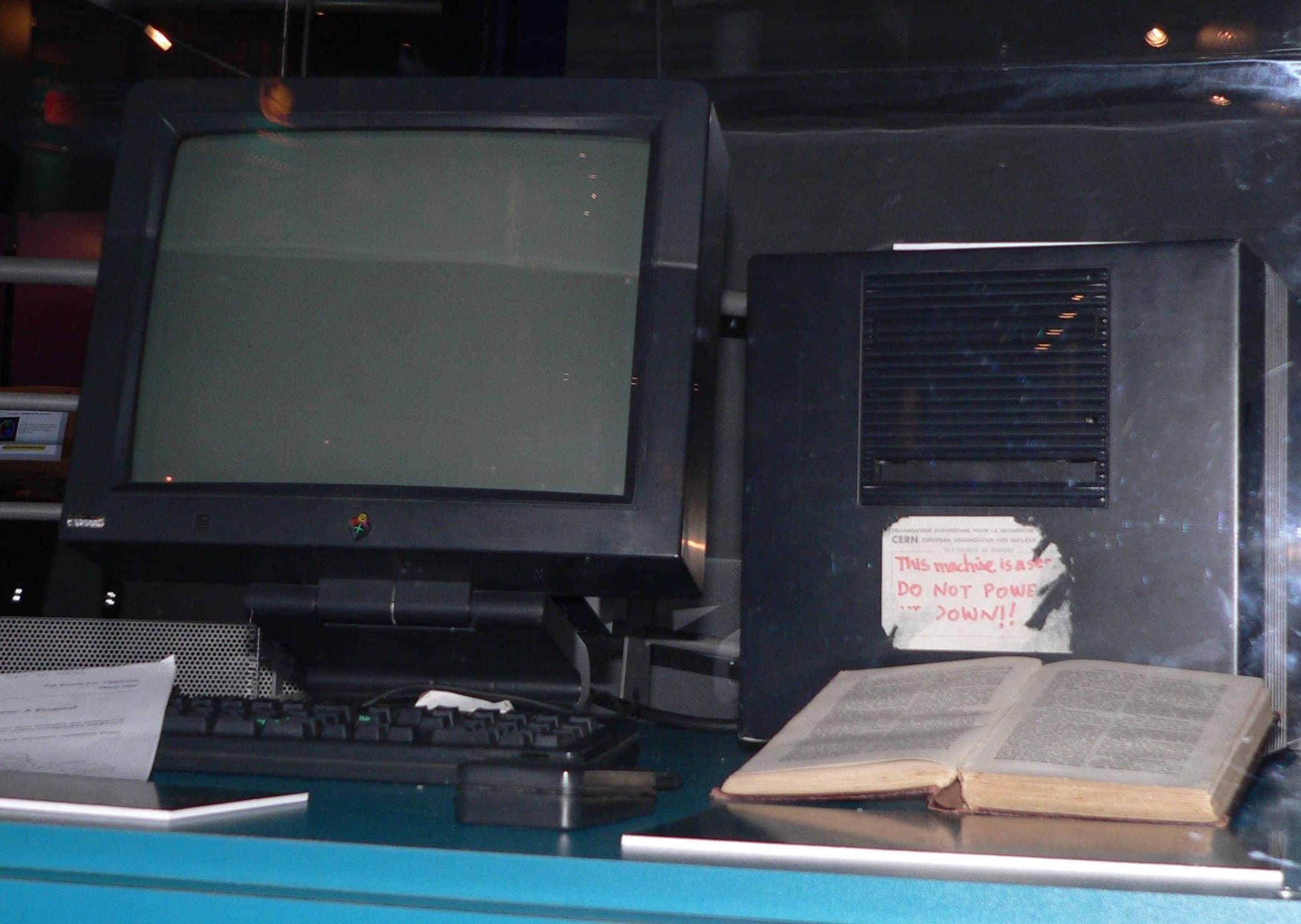 A black NeXT computer with a dark monitor and keyboard in a museum case, next to an open book, with a handwritten note on the machine reading This machine is a server, do not power it down.