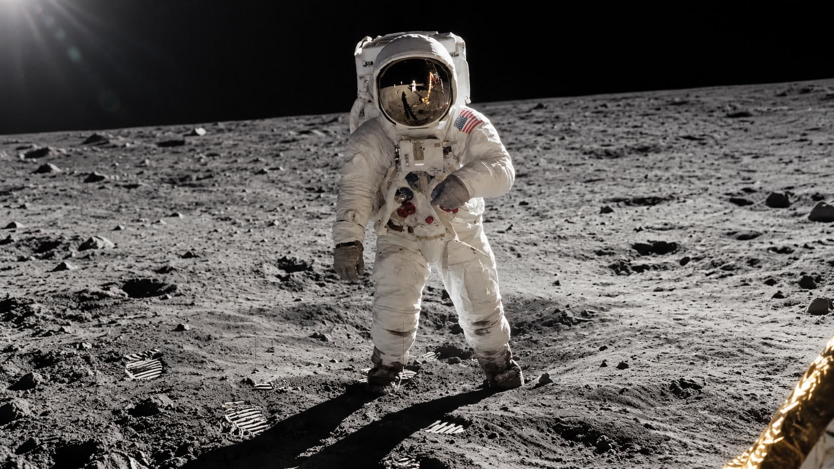 Buzz Aldrin standing on the lunar surface in a white spacesuit, Neil Armstrong reflected in his visor.
