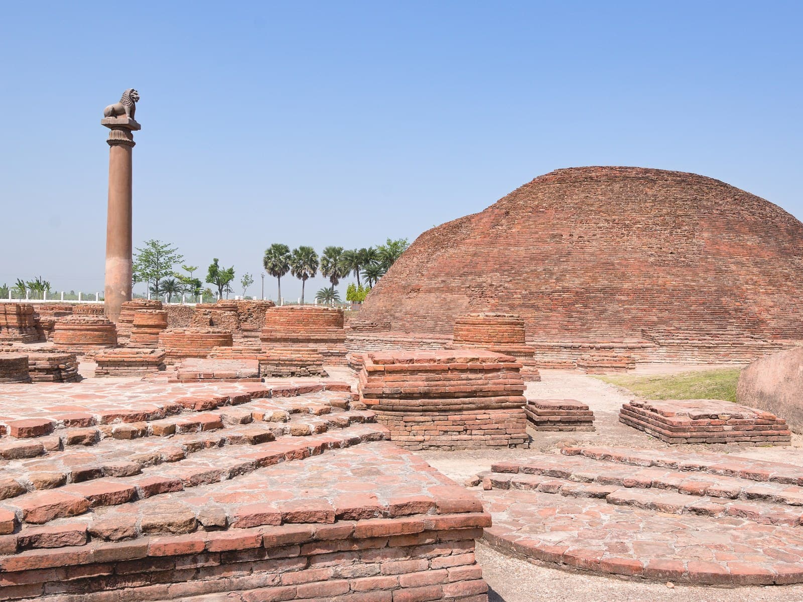 An Ashokan stone pillar standing beside the brick Ananda Stupa at Vaishali, Bihar, under a blue sky.
