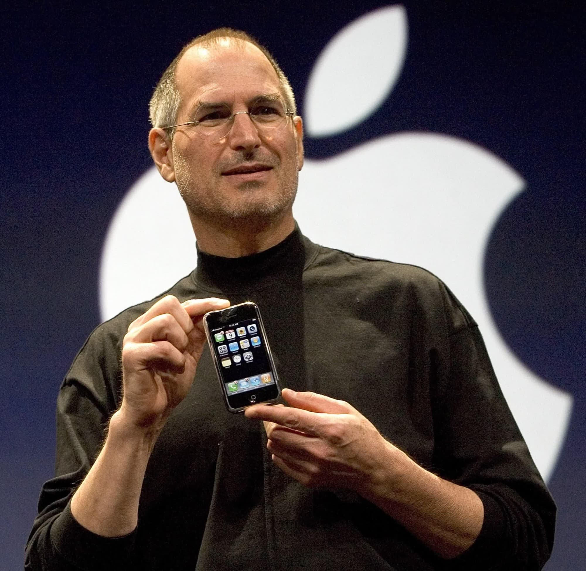 Steve Jobs holding the original iPhone on a stage, the Apple logo on the screen behind him.