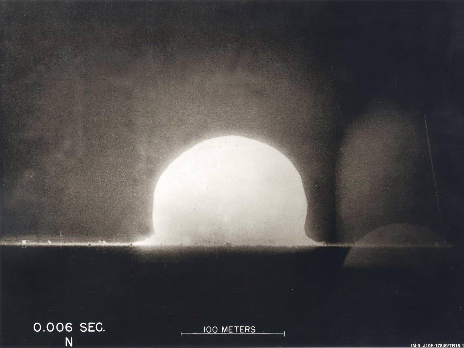 Black-and-white photograph of the Trinity test fireball six milliseconds after detonation, a bright dome above the desert floor, with a 100 metre scale bar.
